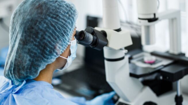 Lab researcher with protective mask peering through microscope