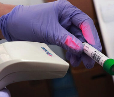 Gloved hand of NYBCe laboratory worker in a testing facility holding a sample of blood in a barcoded vial